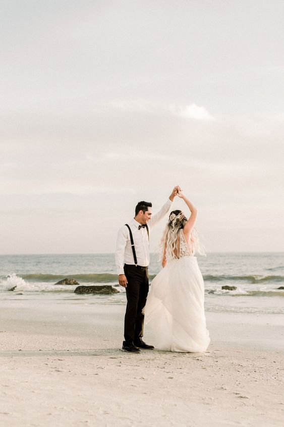Bride and groom twirl in celebration during their celestial themed beach wedding photography shoot. The bride is wearing a long, flowy dress, hair with dark brown to white blonde ombre, while the groom is wearing a modern take on the classic suit - suspenders only, sans jacket. | Celestial Wedding Inspiration with Incredible Star and Moon Details | #equalitymindedweddings #lgbtweddingmagazine #loveinc #beachwedding #bohowedding #floridaweddings